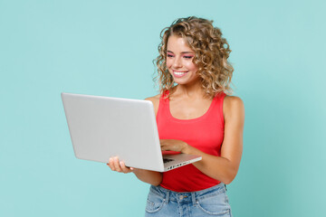 Smiling pretty beautiful attractive young blonde woman 20s wearing pink basic casual tank top standing working on laptop pc computer isolated on blue turquoise colour wall background studio portrait.