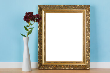 Vertical golden picture frame leaning on blue wall  with stonecrop flowers in vase mockup template. Blank image area isolated with clipping path.