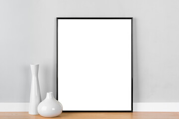 Elegant vertical black picture frame poster artwork mockup template for online shop with white vases in front of light grey wall. Blank image area masked with clipping path.