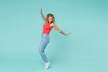 Fototapeta na wymiar Full length side view of excited cheerful young blonde woman 20s in pink tank top jeans standing on toes dancing spreading hands looking down isolated on blue turquoise background, studio portrait.