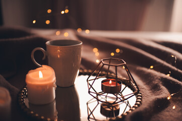 Cup of hot tea with burning candles on tray in bed over Christmas lights close up. Night time atmosphere at home.