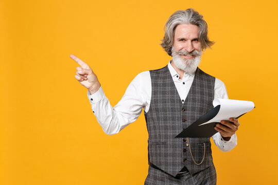 Smiling elderly gray-haired business man in checkered waistcoat shirt hold clipboard papers document pointing finger aside up isolated on yellow background studio. Achievement career wealth concept.