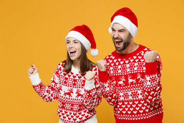Fototapeta premium Overjoyed young Santa couple friends man woman 20s in sweater Christmas hat doing winner gesture clenching fists isolated on yellow background studio. Happy New Year celebration merry holiday concept.
