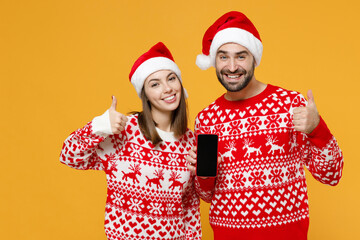 Smiling young Santa couple friends man woman in sweater, Christmas hat hold mobile phone with blank empty screen showing thumbs up isolated on yellow background. Happy New Year celebration concept.