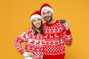 Laughing young Santa couple friends man woman wearing red sweater Christmas hat hold credit bank card isolated on yellow background studio portrait. Happy New Year celebration merry holiday concept.