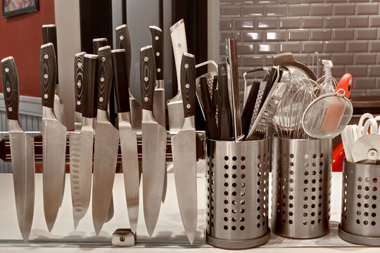 A commercial kitchen worktop, rows of knifes on a magnetic strip and utensils in pots. 