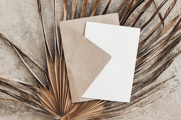Tropical stationery still life. Closeup of blank card mock-up and craft envelope. Dry palm leaf on grunge beige concrete background. Summer vacation concept. Moody boho design. Flat lay, top view.