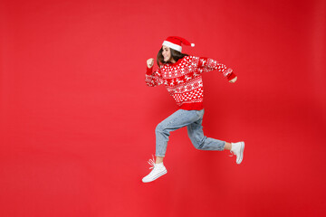 Full length side view of smiling young Santa woman wearing sweater, Christmas hat jumping like running isolated on red background, studio portrait. Happy New Year celebration merry holiday concept.