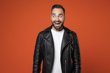 Fototapeta premium Excited surprised funny young bearded man 20s wearing basic white t-shirt, black leather jacket standing keeping mouth open looking aside isolated on bright orange colour background, studio portrait.