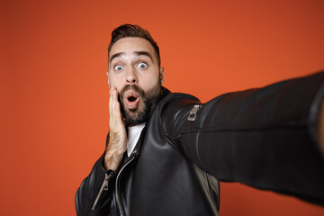 Close up of shocked young bearded man 20s wearing basic white t-shirt black leather jacket standing doing selfie shot on mobile phone put hand on cheek isolated on orange background studio portrait.