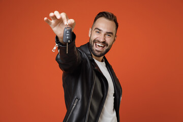 Cheerful successful young bearded man 20s wearing basic white t-shirt, black leather jacket standing hold in hands car keys looking camera isolated on bright orange colour background studio portrait.