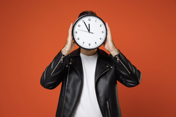 Young man 20s wearing casual basic white t-shirt, black leather jacket standing hold in hands covering face with round clock hiding isolated on bright orange colour background studio portrait.