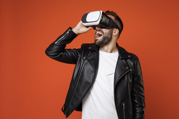 Excited cheerful young bearded man 20s wearing casual basic white t-shirt, black leather jacket standing watching in vr headset gadget isolated on bright orange colour background studio portrait.
