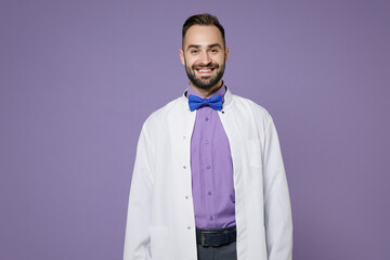Smiling attractive young bearded doctor man wearing white medical gown standing looking camera isolated on violet colour wall background studio portrait. Healthcare personnel health medicine concept.