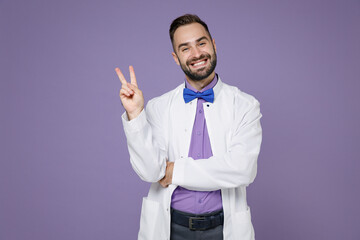 Smiling young bearded doctor man wearing white medical gown showing victory sign looking camera isolated on violet colour wall background studio portrait. Healthcare personnel health medicine concept.