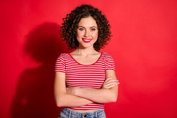 Photo of pretty shiny curly young lady wear casual striped outfit arms crossed isolated red color background