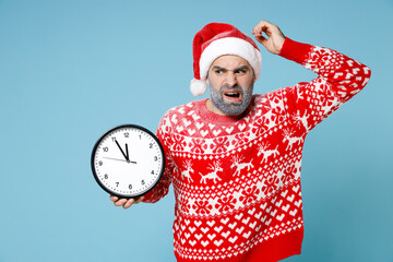 Puzzled young Northern bearded man frozen icy face in Santa hat Christmas sweater hold clock put hand on head isolated on blue background. Happy New Year celebration merry holiday winter time concept.