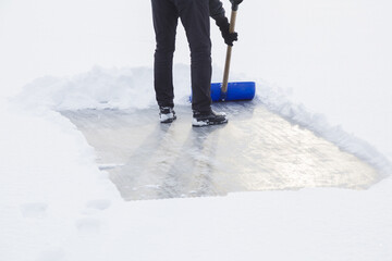 Young adult man cleaning white fresh snow with shovel from ice surface for ice skating. Winter...