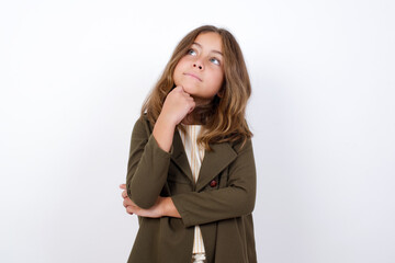Portrait of thoughtful Beautiful little girl standing against white background,  keeps hand under chin, looks away trying to remember something or listens something with interest. Youth concept.