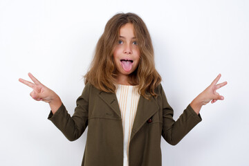 Beautiful little girl standing against white background,  with optimistic smile, showing peace or victory gesture with both hands, looking friendly. V sign.