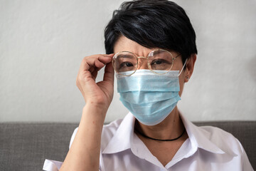 Upset Aged Asian woman with gray hair hand on the glasses is wearing a sanitary mask on her face against coronavirus or COVID-19 disease sitting on a couch at home. And the Elderly concept