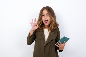 Happy Beautiful little girl standing against white background, sending a message on his smartphone or taking a selfie  and making ok sign with his hand.