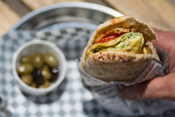 Man hand holding an Omelette sandwich, in a pita bread, with tomato and herbs, served with olives