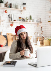 Young brunette woman in santa hat chatting with friends using her laptop at the kitchen