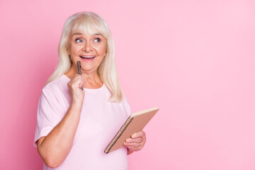 Photo portrait of excited woman touching face cheek with pen holding notepad looking at blank space isolated on pastel pink colored background