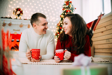 Cute couple on the background of the Christmas tree and the fireplace lying on his stomach with red mugs in his hands