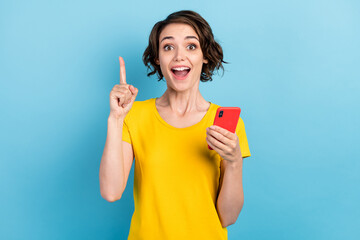 Photo of excited young lady hold smartphone open mouth raise finger wear yellow t-shirt isolated blue color background