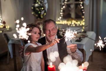 Senior grandfather with small granddaughter indoors at Christmas, having fun with sparklers.