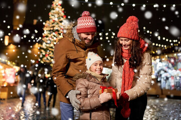 Obraz premium family, winter holidays and celebration concept - happy mother, father and little daughter with gift at christmas market on town hall square in tallinn, estonia over snow