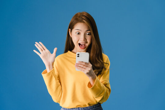 Surprised Young Asia Lady Using Mobile Phone With Positive Expression, Smiles Broadly, Dressed In Casual Clothing And Standing Isolated On Blue Background. Happy Adorable Glad Woman Rejoices Success.