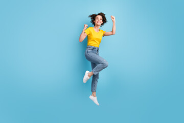 Fototapeta na wymiar Full length photo of funky girl jump raise fists wear yellow t-shirt denim jeans sneakers isolated blue color background