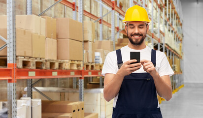 logistics, profession and job concept - happy smiling male worker or loader in yellow helmet and overall with smartphone over warehouse background