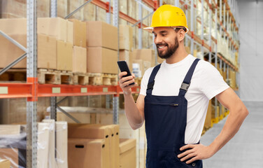 logistics, profession and job concept - happy smiling male worker or loader in yellow helmet and overall with smartphone over warehouse background