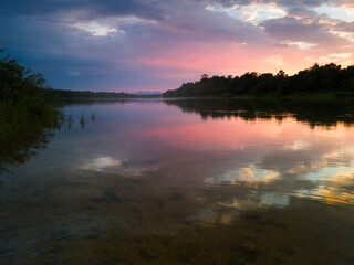 The peaceful landscape of the river and mountain peaks in the distance during a cloudy summer evening, at sunset, the sunlight behind the horizon illuminates the clouds in orange and pink.