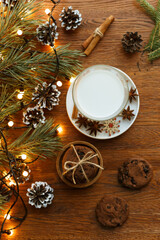 Glass of fresh milk and stack of chocolate cookies arranged on wooden table with pine branches and Christmas garland for Santa 