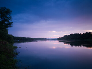 A beautiful summer landscape of a calm river with reflection, gloomy clouds and mystical mountain peaks in the mist in the distance during the blue hour. Weak sunlight colors the clouds bright purple