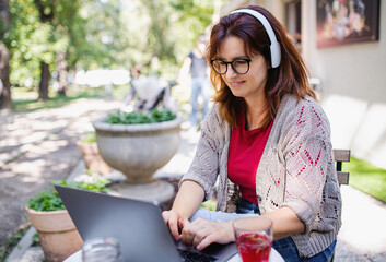 Mature woman with laptop and headphones outdoors in city or town park, working.