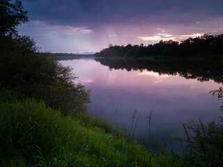Beautiful summer landscape of a calm river with reflection, gloomy clouds with showers and mountain peaks in the distance during the evening. The setting sun turns the clouds purple
