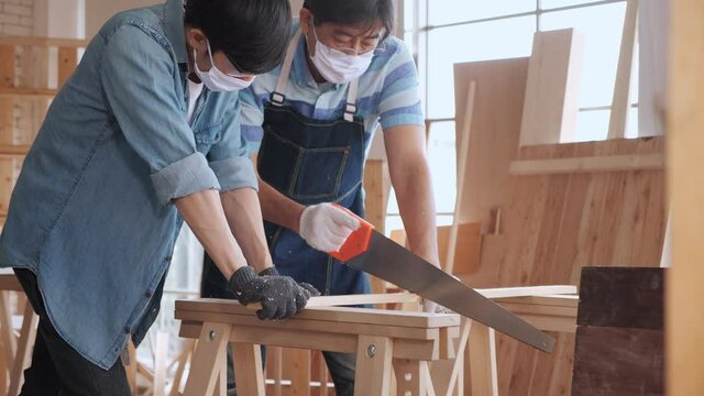 Asian man carpenter training showing apprentice how to use hand sawing in the workshop.