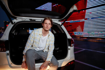 A well-dressed young white long haired guy looking into a camera sitting in an opened car trunk outside on a parking site with a led screen behind him