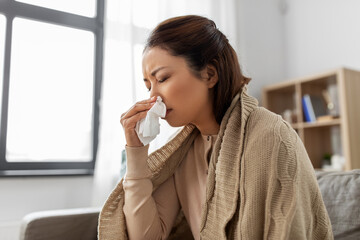 healthcare, cold and people concept - sick asian woman in blanket taking paper tissue from box and blowing her nose at home