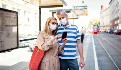 Senior couple with smartphone on bus stop outdoors in city or town, coronavirus concept.
