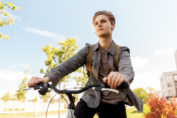 Fototapeta na wymiar lifestyle, transport and people concept - young man or teenage student boy with earphones and backpack riding bicycle in city