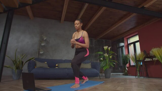 Active sporty fit african american woman doing front forward one leg step lunge exercise for glute and leg muscles, working out watching online fitness training on laptop in domestic interior.