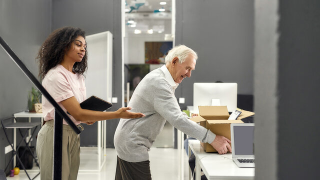 Friendly female company executive supporting, welcoming new employee aged man, senior intern putting cardboard box with belongings on the table - Powered by Adobe