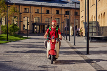 Young cheerful caucasian male courier with thermo bag delivering food to customers, riding scooter...